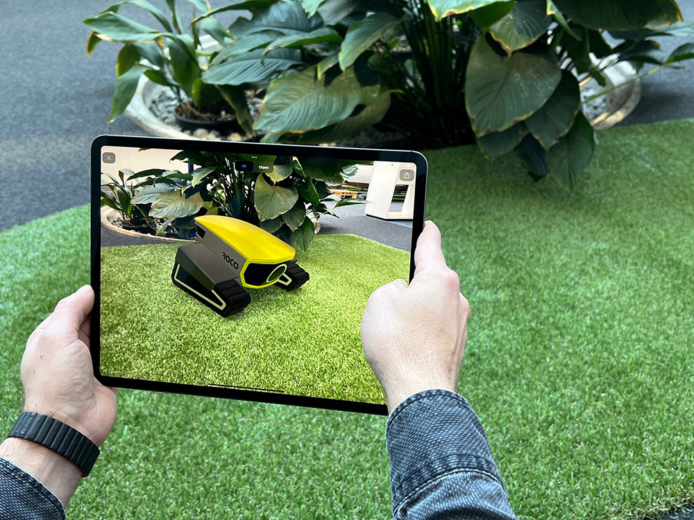 Tablet displaying an augmented reality image of a yellow track crawler on artificial grass next to indoor plants.