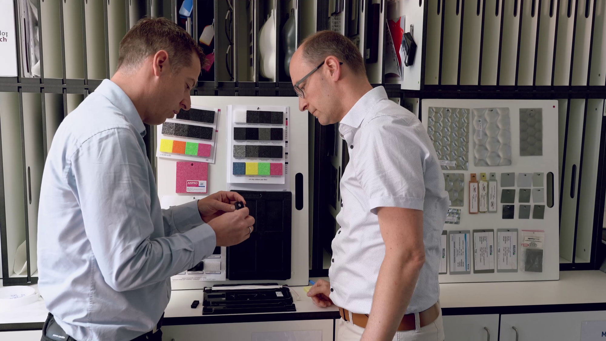 Zwei Ingenieure, die in einem Büro ein 3D-gedrucktes Objekt untersuchen, mit Materialproben im Hintergrund.